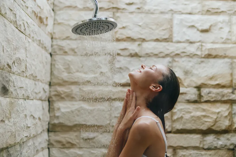 Young woman enjoying outdoor shower with warm water, natural stone wall background, peaceful expression, relaxed moment in daylight.