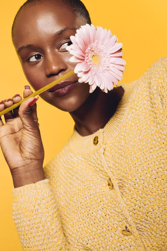 Close-up of a dark skinned African woman holding a pink flower near her face against a yellow background. Concept of beauty, nature, and self-expression with warm lighting.