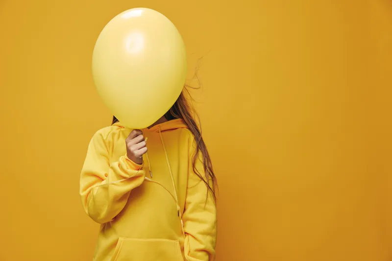 Child in yellow hoodie holding yellow balloon covering face against yellow background for playful and colorful portrait with monochrome style and casual outfit.