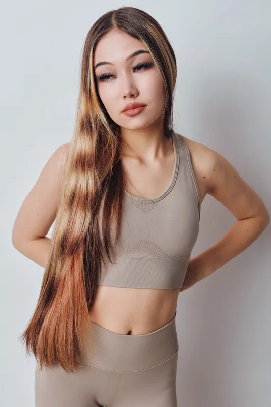 Young woman with long hair wearing beige sportswear standing against white background posing with hands on hips, looking away, lifestyle and fitness concept.