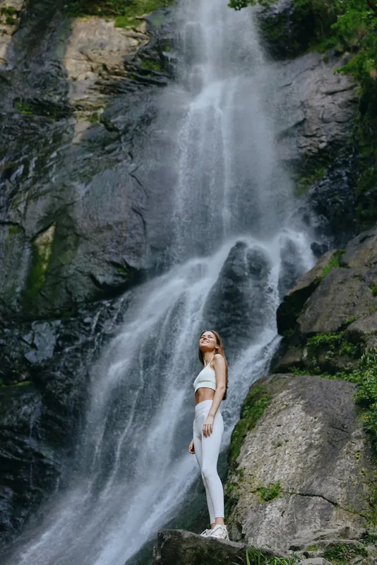 young woman standing near a tall waterfall in white activewear, surrounded by lush green forest and rocky cliffs, enjoying nature and serenity