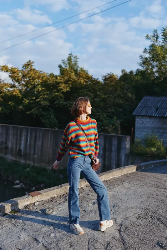 Young woman in a striped sweater and blue jeans stands in a casual outfit on a rural rooftop, small crossbody bag, sideways glance and relaxed smile in golden hour lifestyle travel fashion mood.