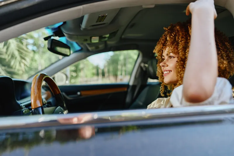 Smiling woman with curly hair sitting inside a car, enjoying a sunny day with natural light and leafy background, radiating happiness and freedom.