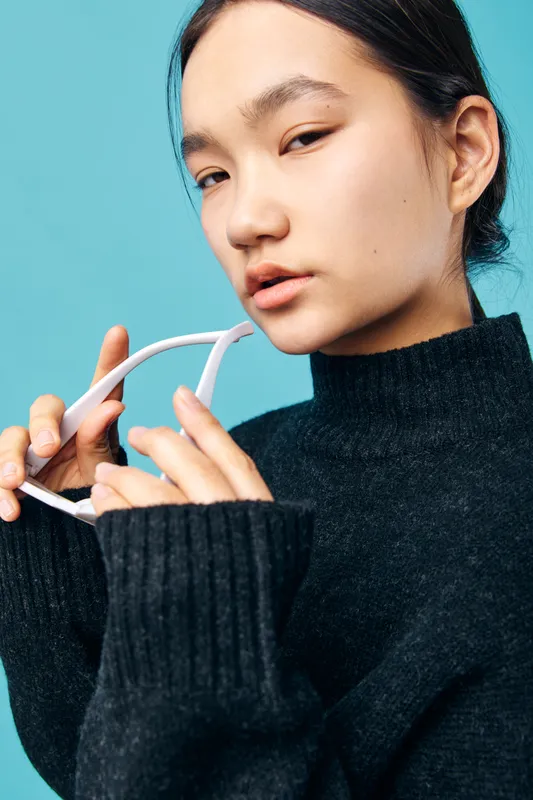 Young woman in black sweater holding white sunglasses near face, posing against blue background with thoughtful expression and natural makeup for fashion portrait.