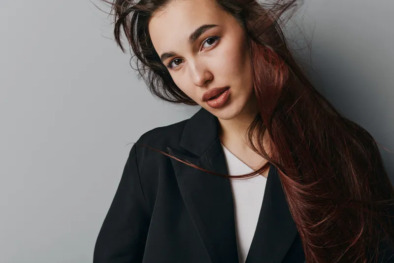 Young woman with long brown hair and natural makeup wearing black coat and white top posing against gray background with confident expression and casual hairstyle.