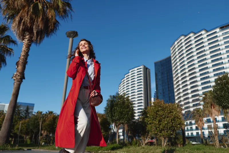 Young woman in a red trench coat walks confidently in an urban park while talking on her smartphone on a sunny day with modern buildings in the background.