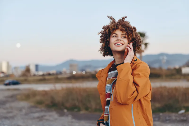 Smiling woman with curly hair in an orange jacket, outdoors by a road path, hand near face, looking at camera, lifestyle portrait in autumn scenery, soft evening light