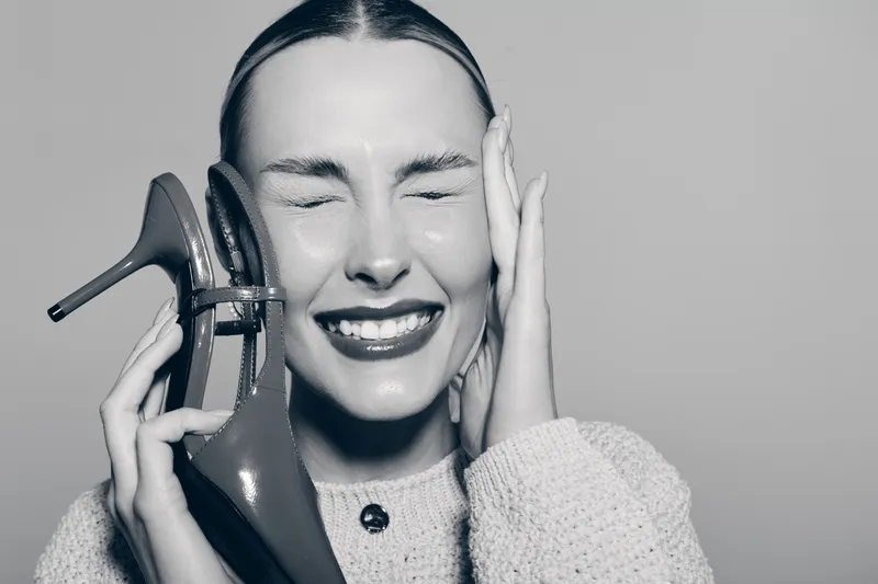 Young white woman with light skin holds high heel shoe to her ear as if it is a phone, eyes closed and smiling. Concept for humor, fashion, and playful emotions in studio.