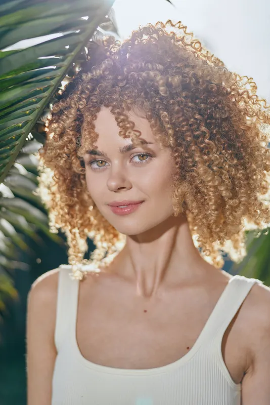 smiling woman with curly hair in white top stands near tropical leaves under sunlight, natural beauty, warm summer day, outdoor portrait