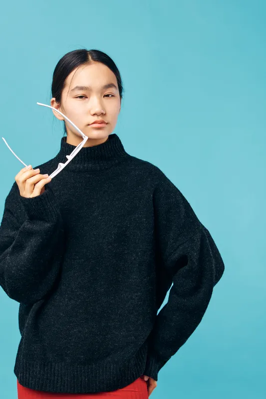 Young woman in black sweater holding white sunglasses with confident expression against blue background. Female model with casual style and relaxed pose for fashion portrait.