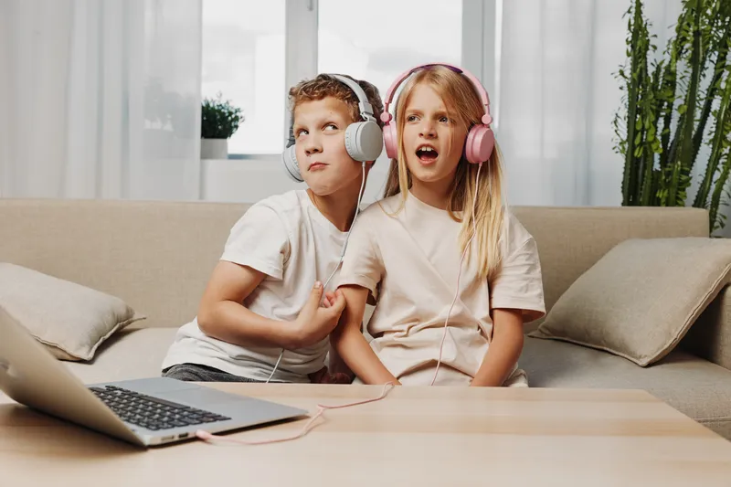 Two kids with headphones share music on couch, enjoying playful moment with laptop in cozy living room. Concept of childhood, technology, and entertainment.