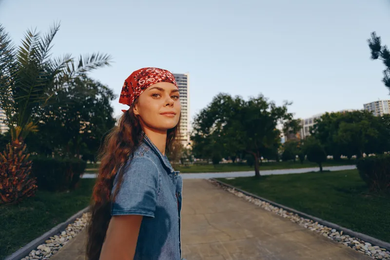 Lifestyle woman in modern boho-western denim street style wearing a red bandana, walking confidently outdoors with a warm film color and natural background.