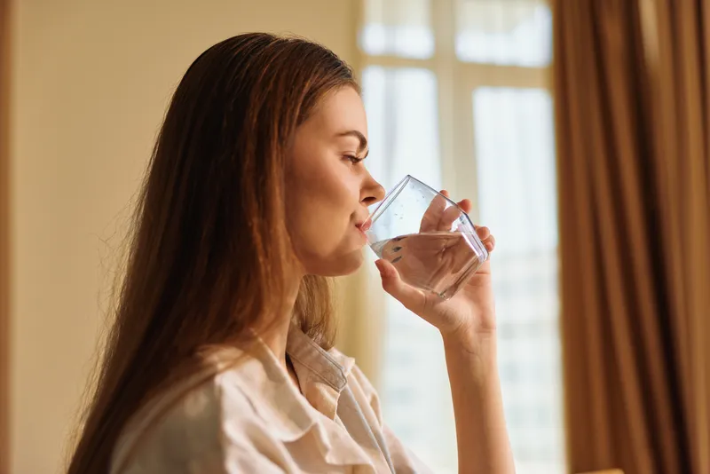 Woman drinking water from glass in profile at home morning, healthy hydration portrait with natural window light, casual longhair female sipping in relaxed domestic routine.