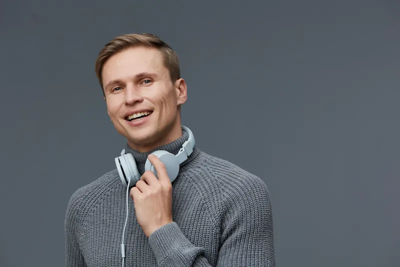 Smiling man wearing gray sweater and headphones on neck, holding hand near face, looking at camera, friendly expression, studio portrait with plain gray background, casual style, happiness, technology