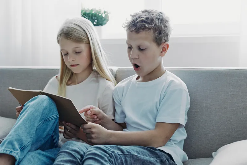 Two children sitting on sofa reading digital tablets together, focused and engaged in technology learning at home in bright natural light.