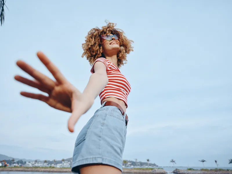Young person wearing striped crop top and denim shorts stretches toward camera on a sunny day by the sea, creating a dynamic, inviting sense of freedom and motion.