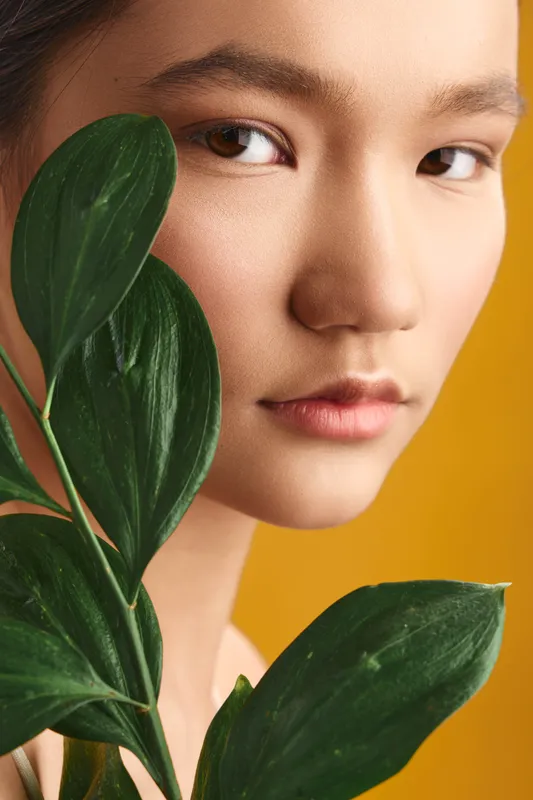 Asian woman gazes softly at the camera while leafy plants frame her face, set against a warm yellow backdrop, conveying calm, natural beauty and minimalist elegance suitable for lifestyle and