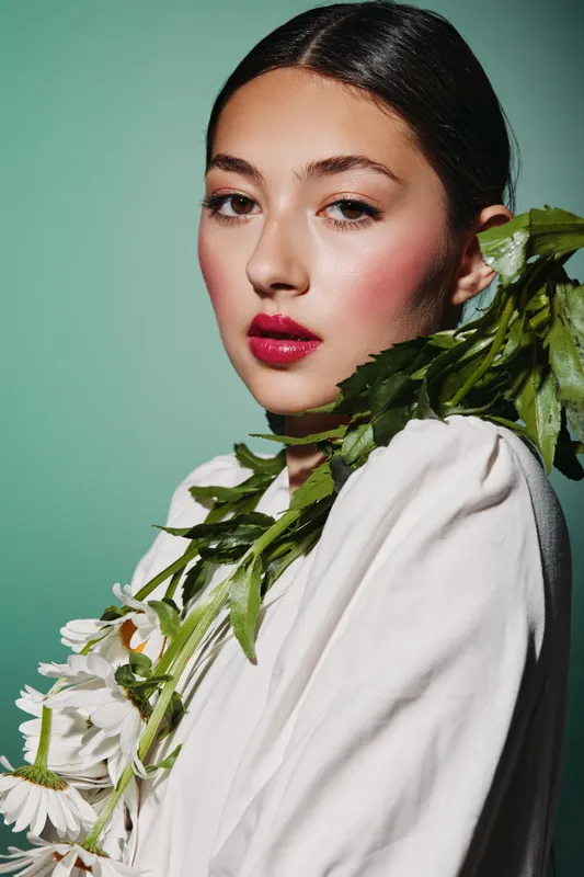 Portrait of a young woman with glossy red lips and subtle eye makeup wearing a white blouse and greenery wrapped around her shoulder. Beauty fashion concept with creative styling.