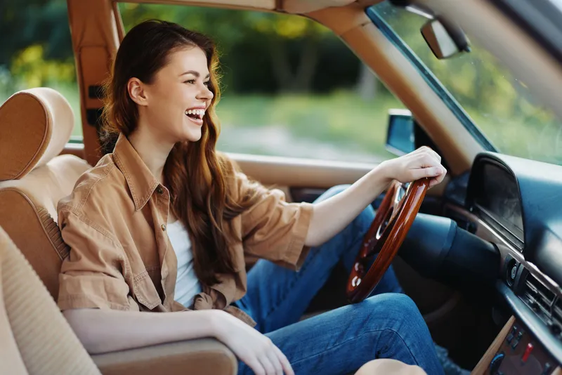 Young woman joyfully driving a vintage car, showcasing carefree emotions in a natural green setting, perfect for summer travel and adventures.