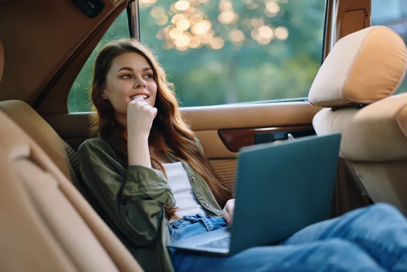 Young woman working on laptop in a car, showcasing a relaxed atmosphere with a sunny background, casual clothing, and a pensive expression.