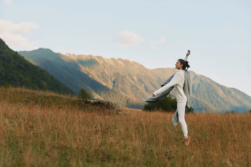 Female figure in a white jumpsuit dances across a sunlit meadow, mountains distant, conveying freedom, motion, and carefree outdoor living with a breeze and open sky.