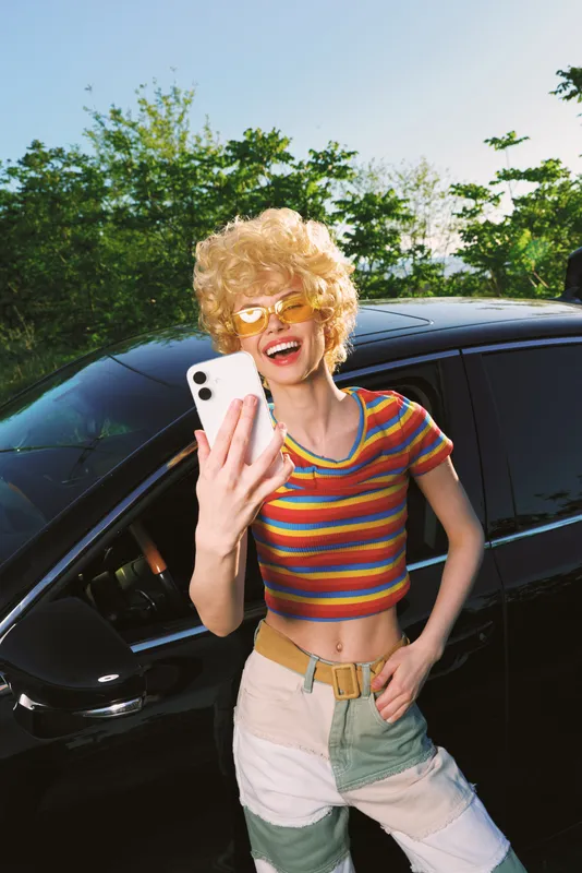 A woman with curly hair in a striped crop top poses by a sleek car, taking a selfie in bright sun. Retro film color tones and colorful streetstyle convey a carefree lifestyle.