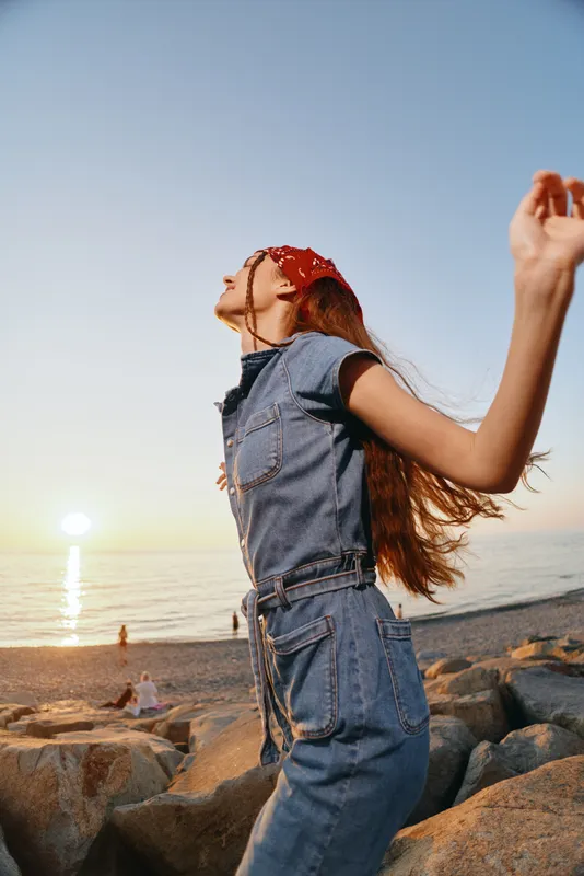 Lifestyle woman in modern boho-western denim street style enjoys sunset on rocky beach with relaxed mood and film color tones, capturing carefree summer freedom.