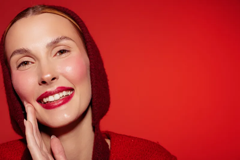 Smiling woman with red lips and rosy cheeks touches her face gently. She wears a red hooded sweater against a bright red background. Concept of beauty, skincare, and happiness.