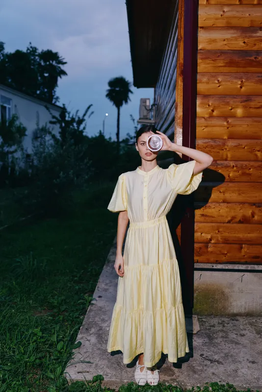Girl in a yellow dress stands beside a wooden house at dusk, peering through a magnifying glass with curiosity, creating a whimsical, investigative mood.