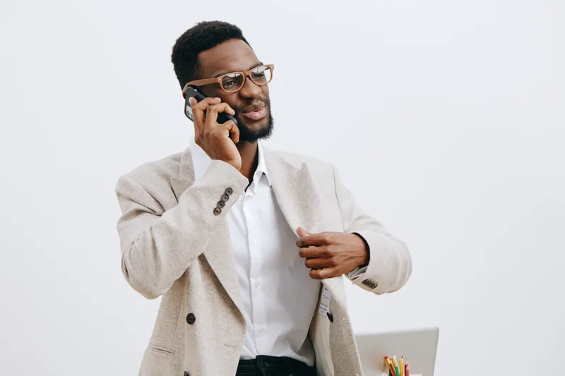Professional young African-American man in smart casual attire talking on mobile phone while holding a pen, standing against a plain white background. Studio portrait, business lifestyle concept.
