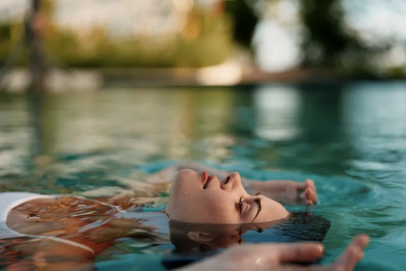 Relaxed woman floats in a pool, eyes closed, enjoying relaxation and leisure under sunshine, with serene calm water and reflections creating a peaceful summer mood.