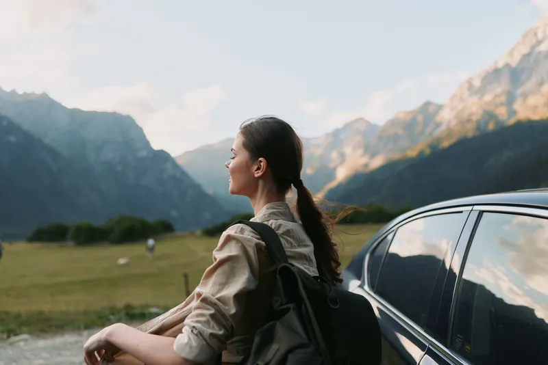 Woman traveler rests beside a car in a mountain valley, backpack on her shoulders, enjoying fresh air and distant peaks, conveying calm independence and outdoor lifestyle