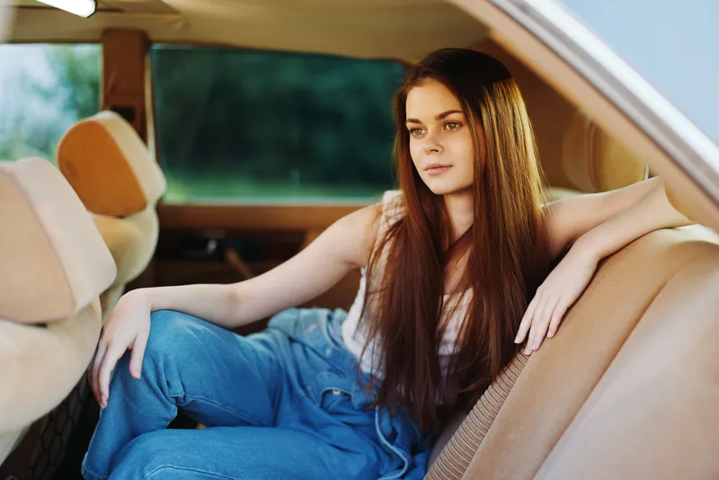 A young woman in casual clothing relaxes inside a classic car, reflecting a carefree spirit and summer vibes with her long hair and thoughtful expression.
