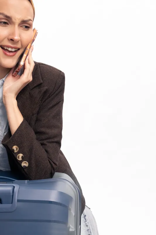 Young woman in brown jacket sitting on suitcase holds phone to ear with worried expression on white background. Concept for travel stress, communication, and urgency.