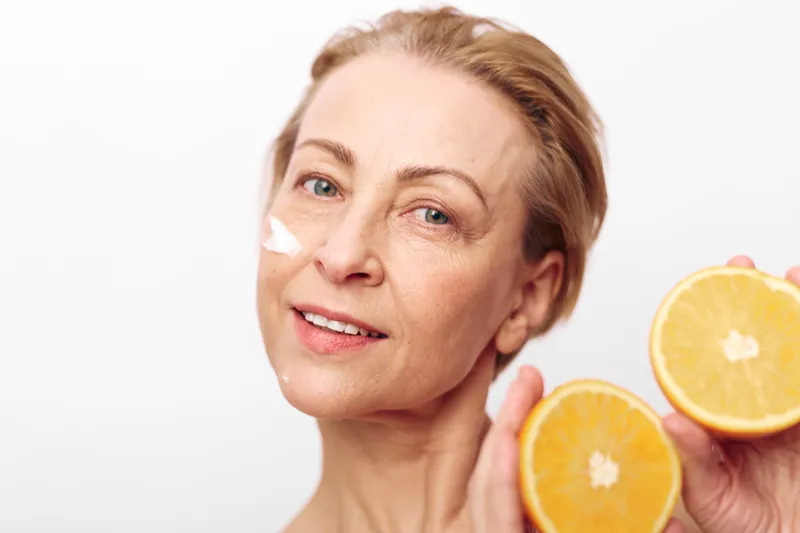 Mature woman with natural skin holds two orange halves near face, applying white cream on cheek. Bright studio light on white background. Concept for skincare, vitamin c, and healthy glow.