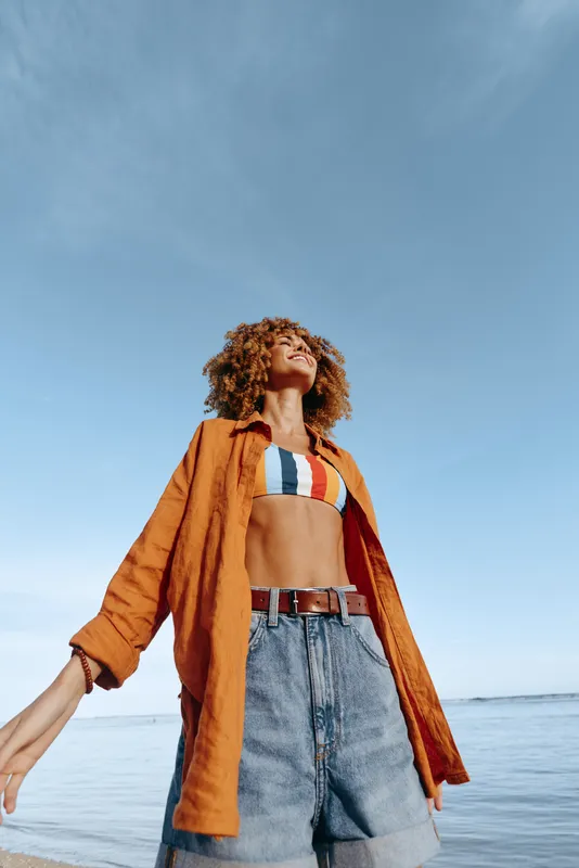 Vibrant woman on a beach wearing an orange shirt over a striped bikini and denim shorts