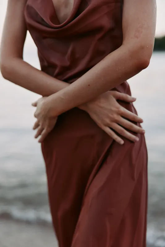 Close-up of a person wearing a burgundy dress, arms crossed at the waist, fabric draping softly. Minimal waterfront background highlights form, texture, and quiet elegance.
