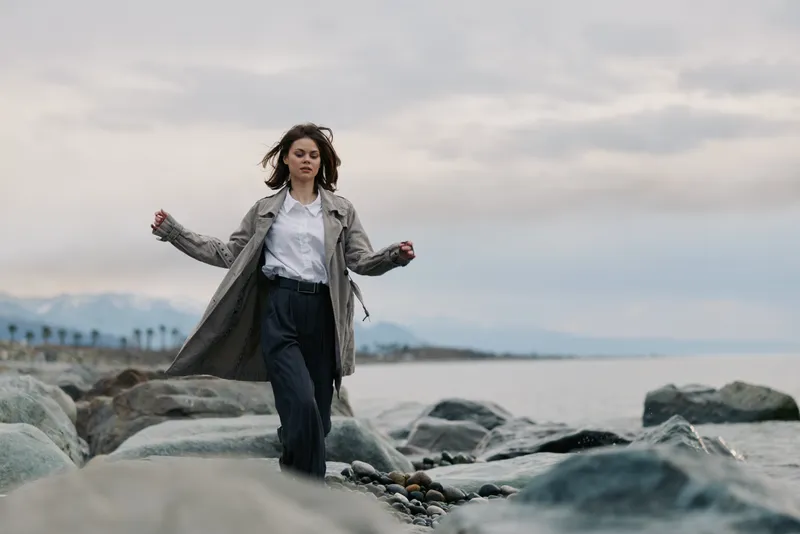 Young woman wearing long coat walks on rocky shore near calm sea under cloudy sky. Concept of autumn fashion, outdoor lifestyle, and serene coastal atmosphere with natural background.