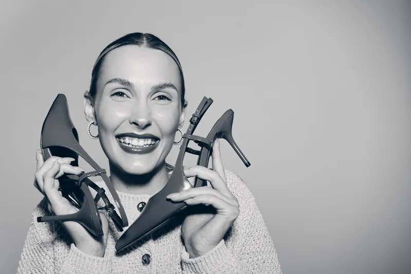 Smiling white woman with fair skin holds high heel shoes near face, wearing cozy sweater. Concept for fashion, happiness, and style.
