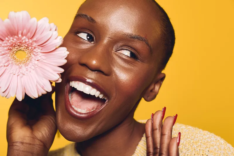 Happy African woman with dark skin holding pink flower near face, smiling with open mouth on yellow background. Concept for joy, beauty, skincare, and natural cosmetics.
