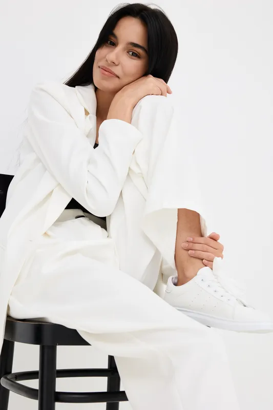 Woman in white suit sitting on black chair with relaxed smile, dark hair, casual pose, minimalistic bright background, stylish and modern look.