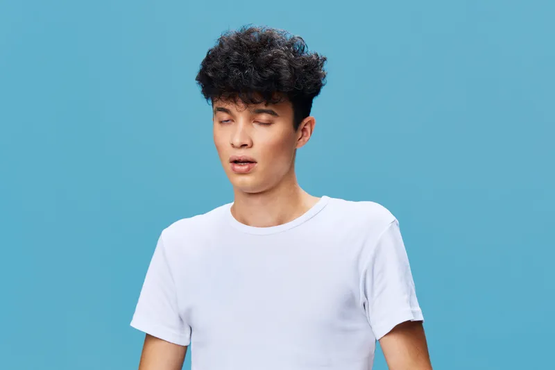 young man in white t-shirt with curly hair grimacing with closed eyes against solid blue background, expressing discomfort or unpleasant sensation, casual style