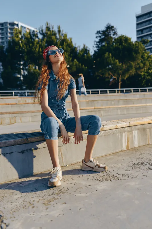 Lifestyle woman in modern boho-western denim street style sitting outdoors in casual outfit with sunglasses, relaxed pose, and urban sunny background in warm film color.