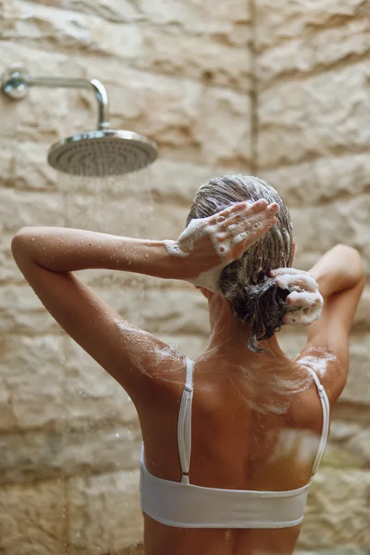 Woman washing hair under shower with soap foam on skin and wet hair, relaxing in modern bathroom with natural stone wall background and warm lighting.