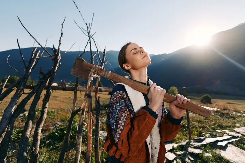 Woman with axe on shoulder in countryside field with fence and mountains, sunlight bathing her face; rural portrait of outdoor work, nature, farming lifestyle and peaceful relaxation.