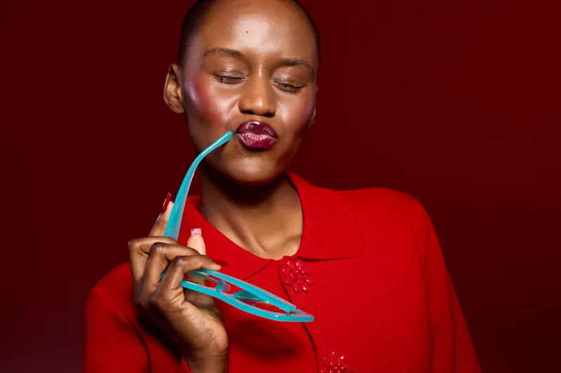 Portrait of a dark skinned African woman in red clothing holding blue sunglasses, lips pursed. Studio shot with red background. Concept of fashion, beauty, and confident expression.