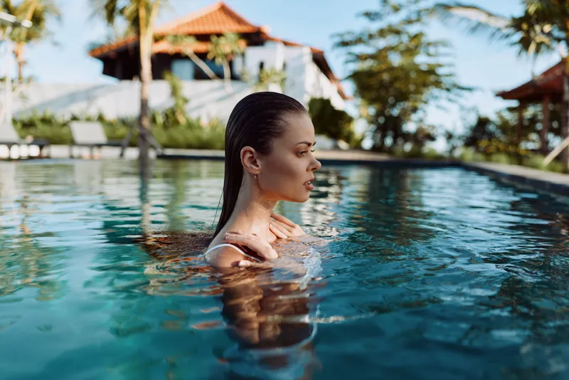 Woman stands in a clear pool at a tropical resort, calm expression, blue water surrounds her, palm trees in the background, leisure lifestyle and vacation mood, outdoors and serene.