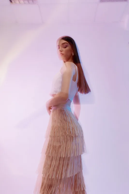 Young woman in a stylish beige fringe skirt and white top poses against a soft purple background. Concept for fashion, beauty, elegance, and modern style.