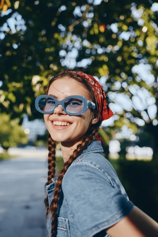 smiling woman with blue sunglasses wearing denim jacket and red headscarf outside on sunny day with green trees in background