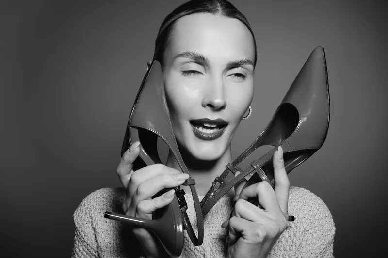 Young white woman with light skin holds high heel shoes near face with eyes closed and playful expression on dark background. Concept of fashion, style, and footwear.